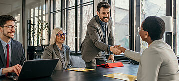 Vier Personen in einem modernen Büro, ein lächelnder Man im Anzug, schüttelt einer ihm gegenüber sitzenden Frau die Hand.