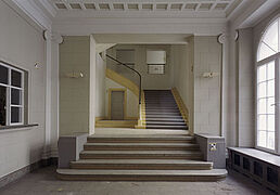 Blick in einen großen, von Tageslicht erhellten Raum von dem eine breite Treppe in ein dahinter anschließendes Treppenhaus überleitet.