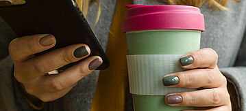 Eine Frau hält in einer Hand ihr Smartphone und in der anderen einen Mehrwegbecher
