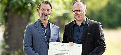 zwei Männer stehen im Wald mit einem Förderbescheid