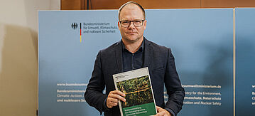 Carsten Schneider hält den ANK-Vorschlag des BMUKN als Broschüre in die Kamera